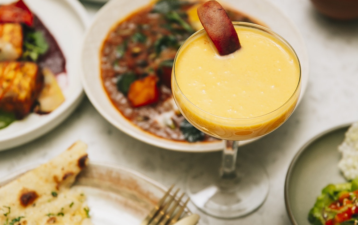 A mango lassi cocktail on a table surrounded by Indian share dishes
