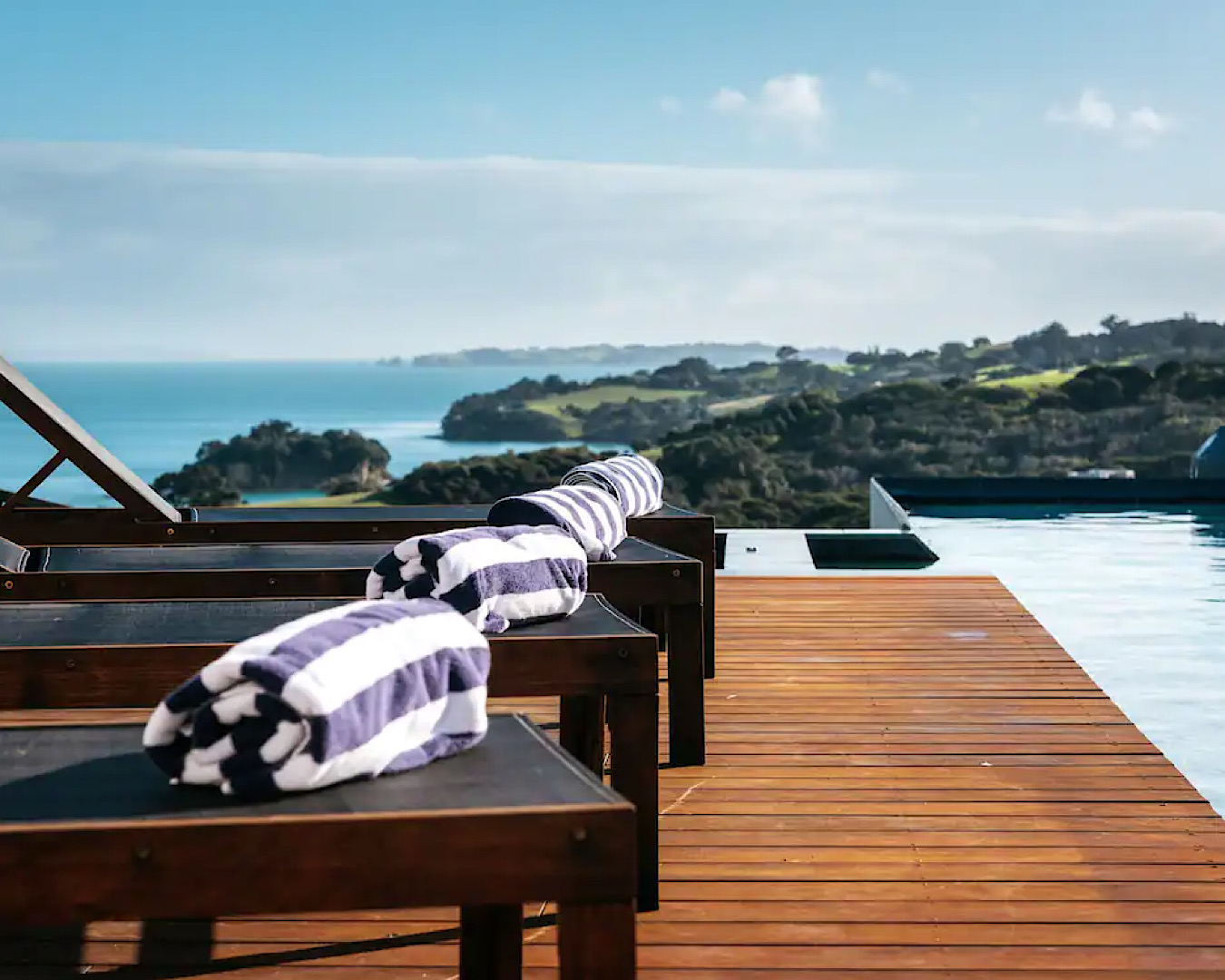 A stunning pool and row of deck chairs with stripy towels on them overlooking an incredible view of the Hauraki Gulf. 