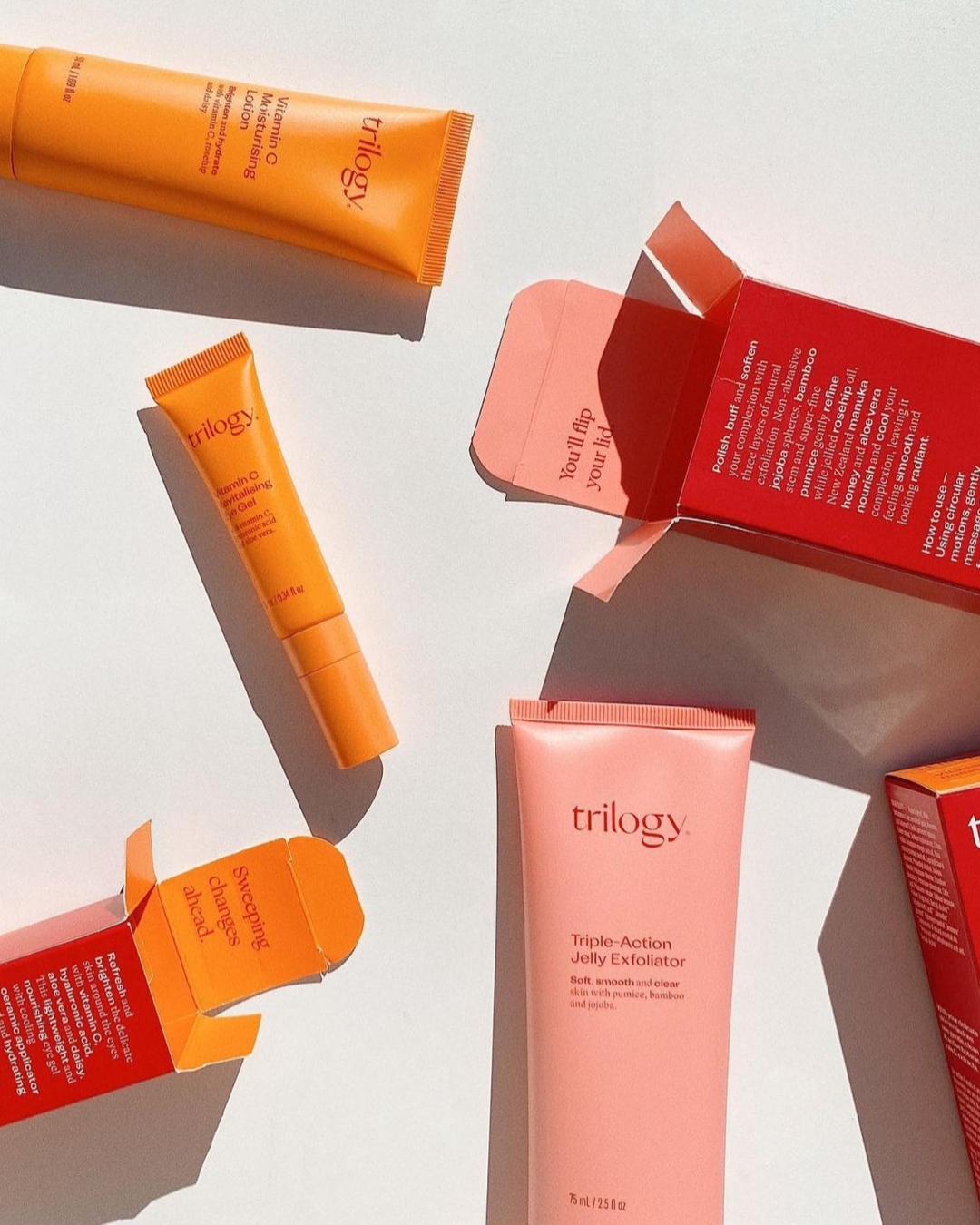 Orange and pink tubes of Trilogy skincare and red boxes artfully spread out on a white backdrop.