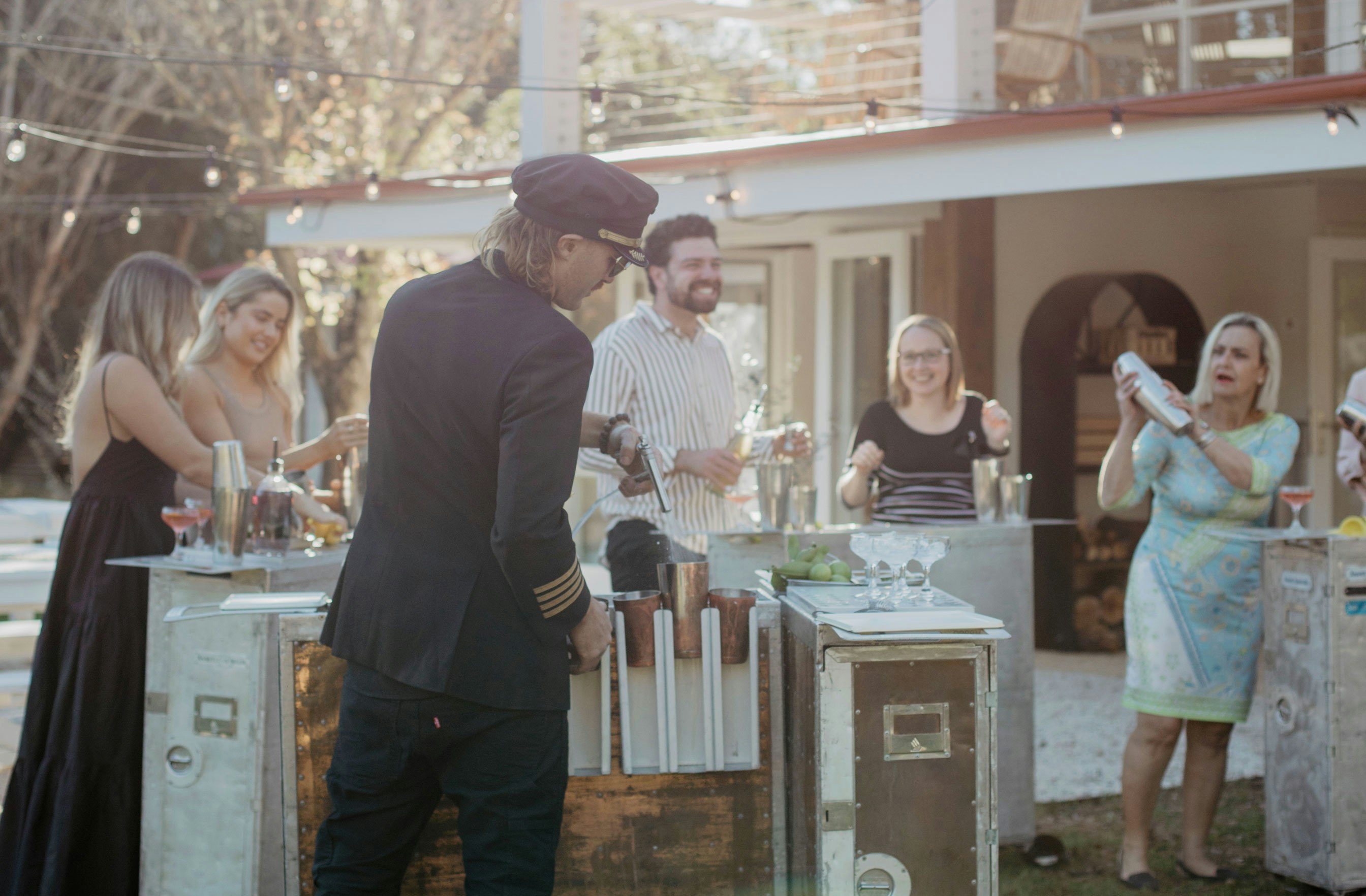 Man with back to camera prepares cocktails for 5 guests at an outdoor garden party