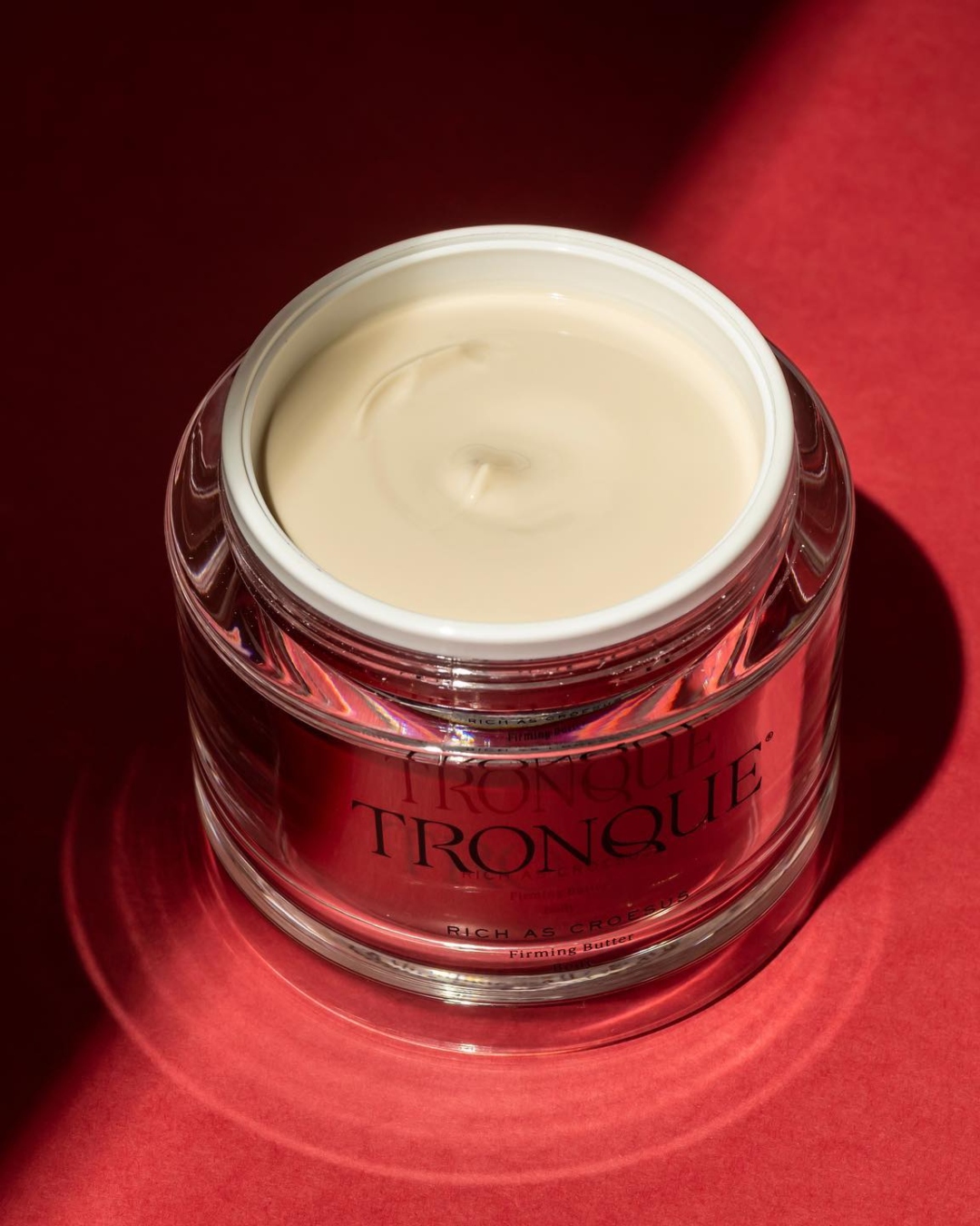 A red glass tub of Tronque firming butter on a sunlit red backdrop.