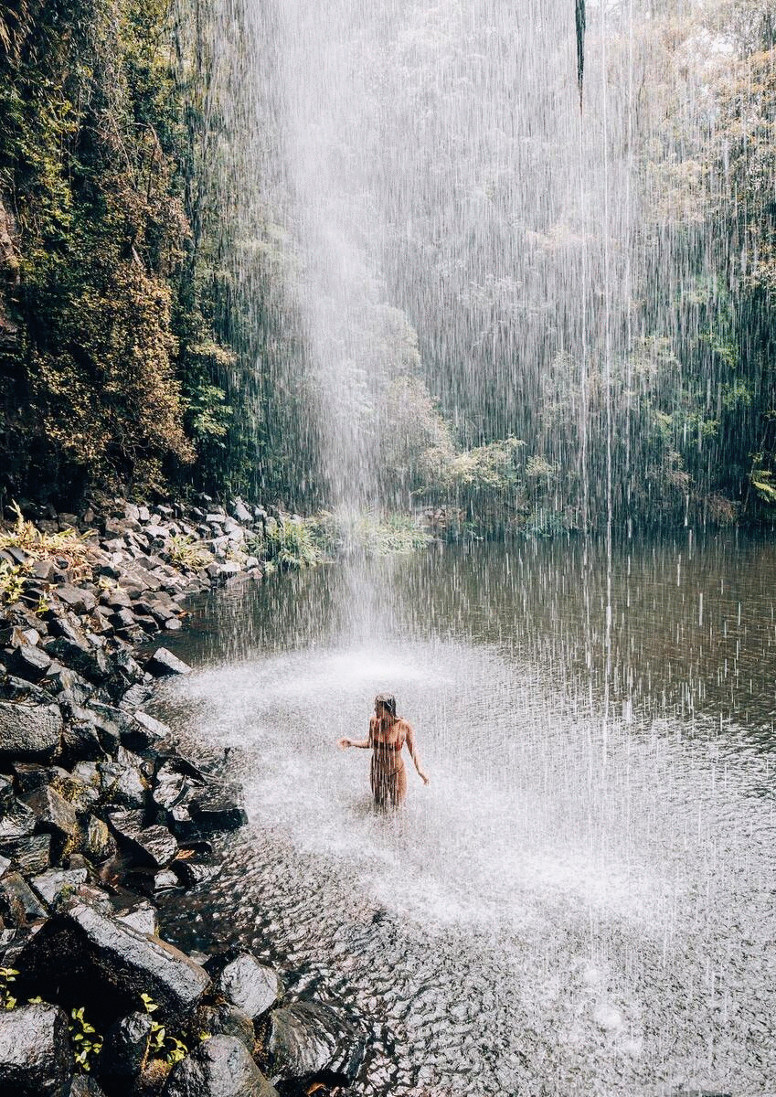 A woman stands under a waterfall surrounded by rainforest.