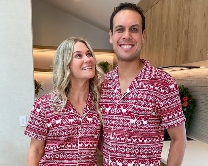 A couple stand together wearing matching red pyjamas inspired by ugly Christmas sweaters. 