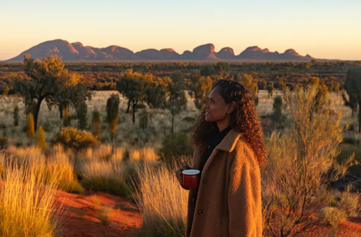 A woman sips a cup of coffee with Uluru in the background at sunrise