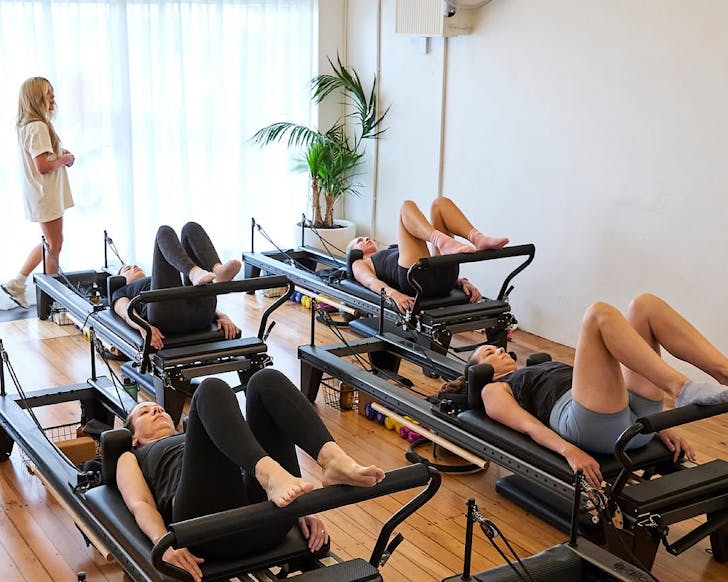 A teacher in a white t-shirt dress roams a studio while participants lie on their backs on reformers with their heels on the foot bar. 