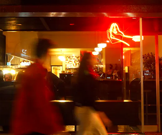 Two people walking past a shop front with a neon lit hand gesturing in Italian. 