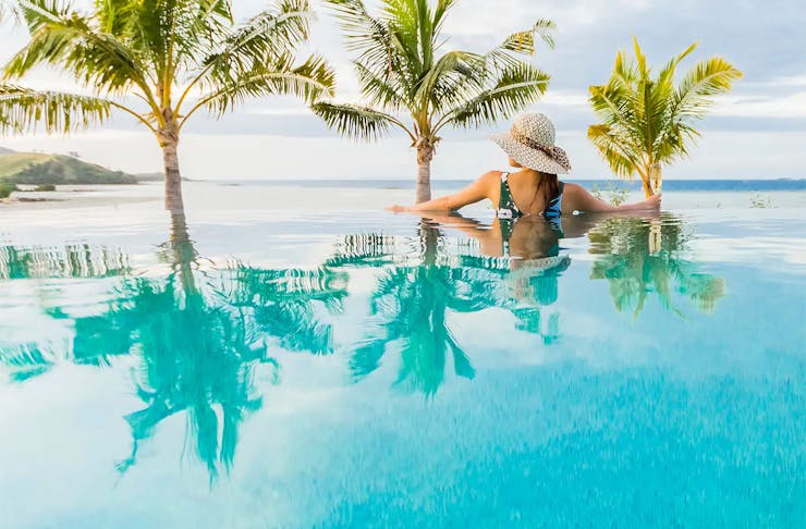 A woman wearing a green and white swimsuit and a straw hat looks out to sea while soaking in an infinity pool at Vale-I-Yata that appears to merge seamlessly with the ocean. 