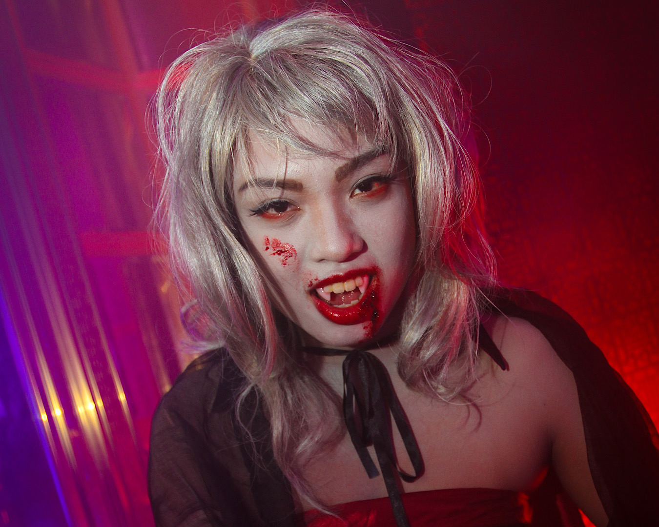 A girl dressed as a vampire in a pink purple neon room. She wears a silver wig, fangs, a cape and has blood smeared down her her cheek and chin. 