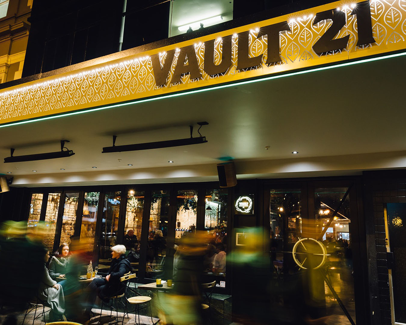 People sit outside Vault 21 which is lit up with a yellow hue.