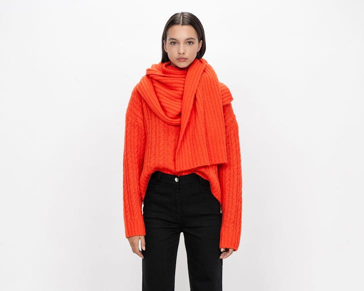 A woman wears a large cable knit orange jumper and scarf. 