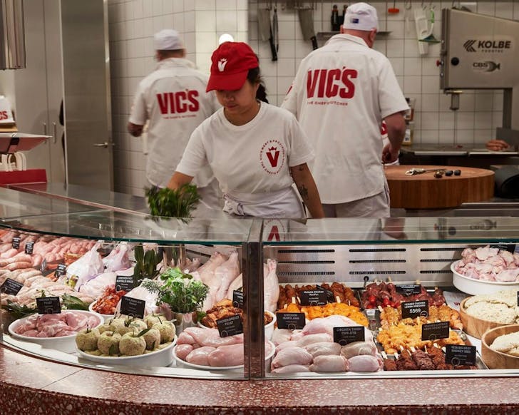 A butcher window full of meats with three butcher's working inside the store, with shirts that read 