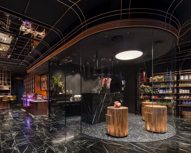 A wide shot of the interior of the Victor Churchill butcher. The floor is sleek green marble and the colours of the interior are dark. There are walls of butcher grocery products to the right and seating to the far left back corner. In the centre and main stage of the image is a display meat cutting area, with block wooden tables connected to the ground where butchers chop the meat. 
