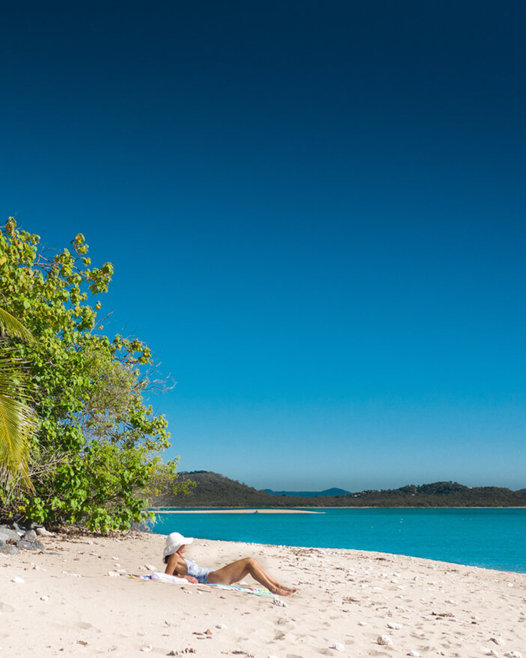 A woman sunbakes on a white sandy beach on Victor Island overlooking clear blue water.