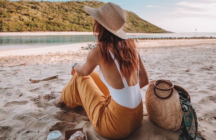 A woman sits on a white sandy beach next to a coffee and croissant. 