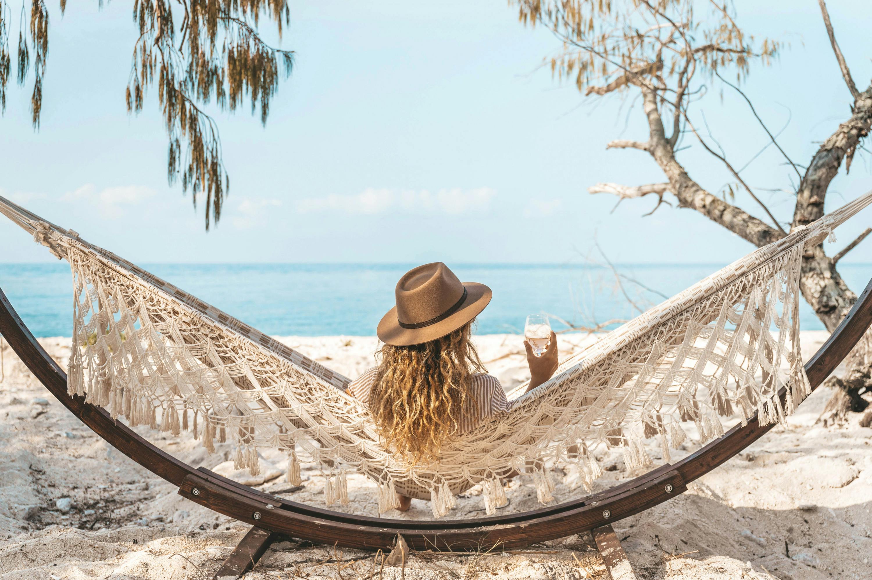 A woman sits on the beach in a hammock with a wine in hand.