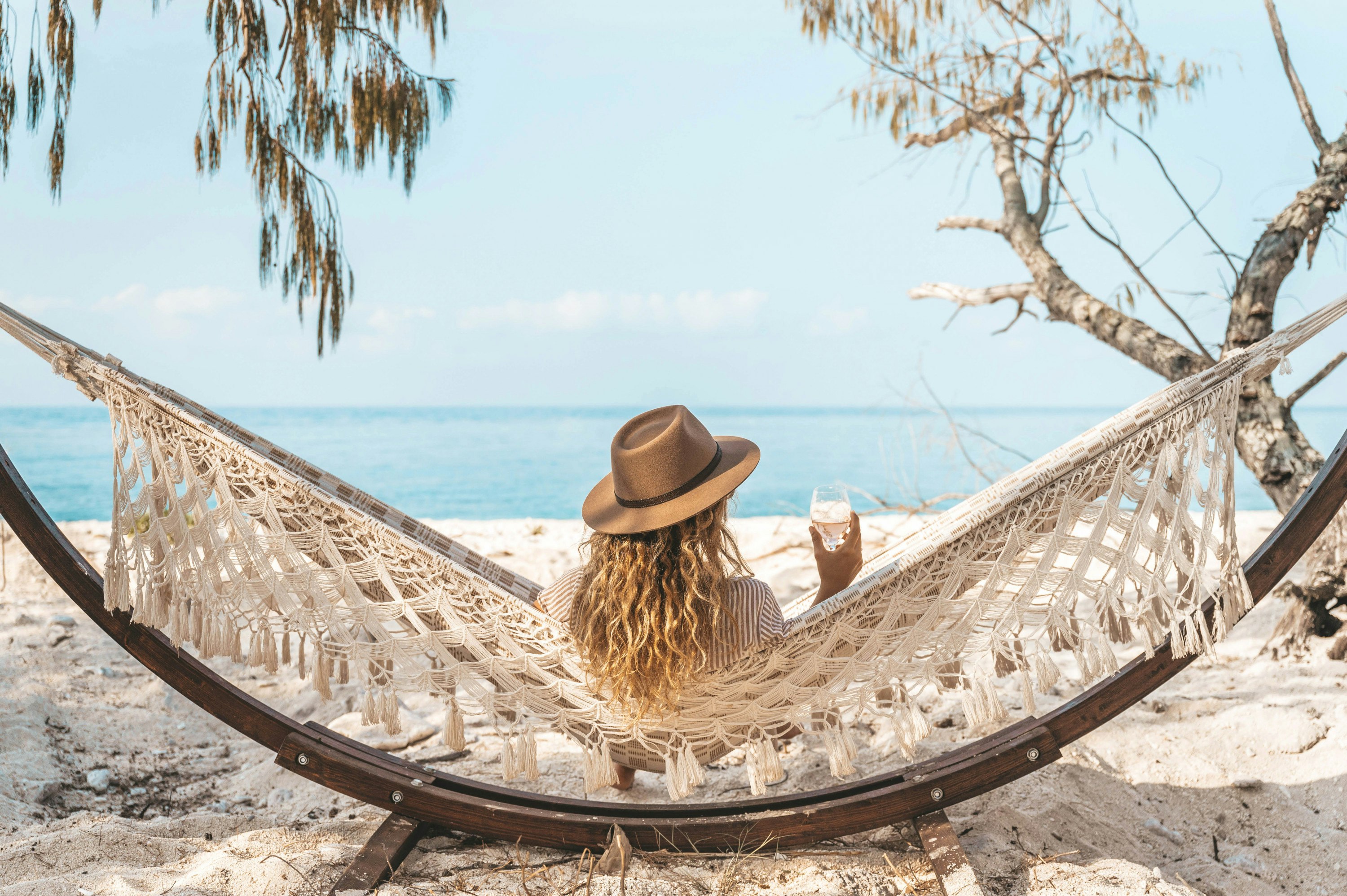 A woman sits on the beach in a hammock with a wine in hand.