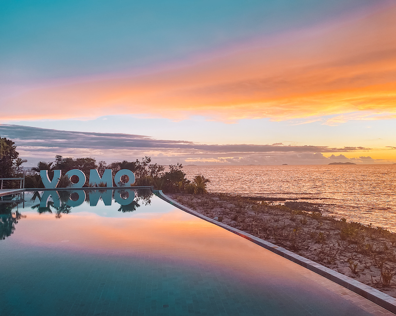 The sun sets over a beautiful pool at Vomo Island Resort. Large white letters spell ‘V-O-M-O’. 