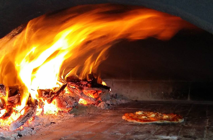 A pizza cooks next to big open flame in a wood fire pizza oven 