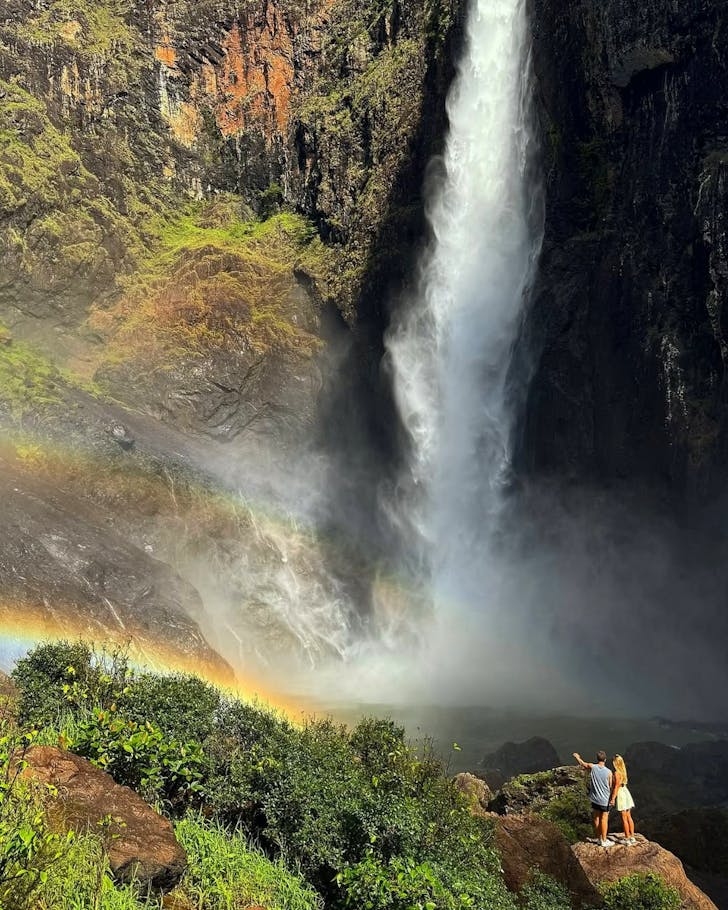 A couple stand together on a rock looking up at Wallaman Falls, a rainbow passing through the mist.