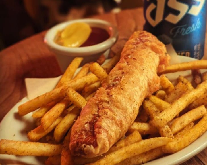 A crispy beer-battered hot dog on fries with two-tone sauce and a can of Cass beer. 