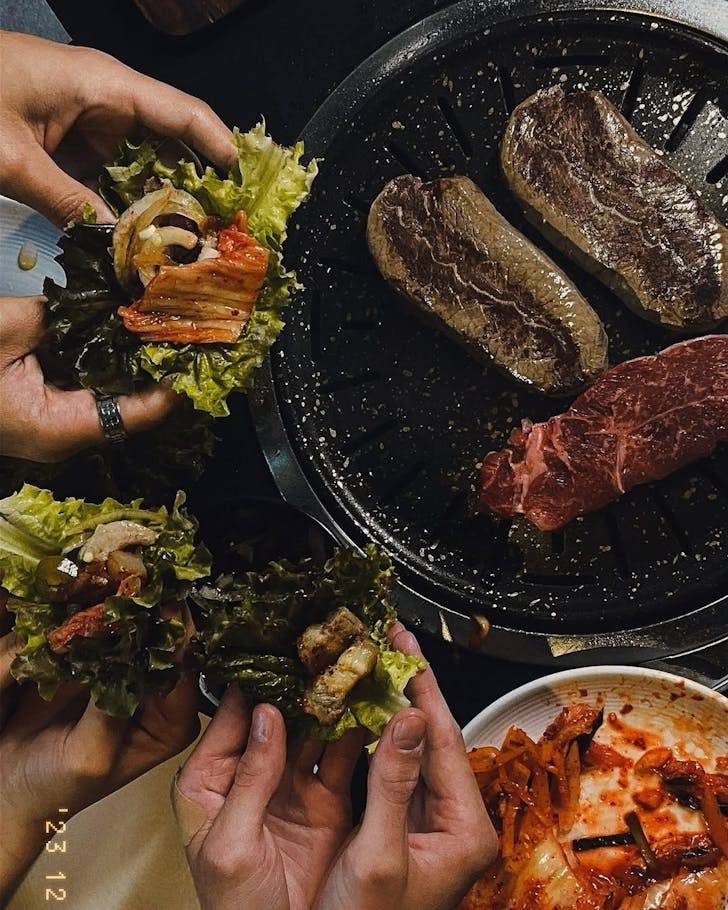 Hands holding lettuce wraps with Korean BBQ meat filling, with steaks grilling and a dish of kimchi beside them.