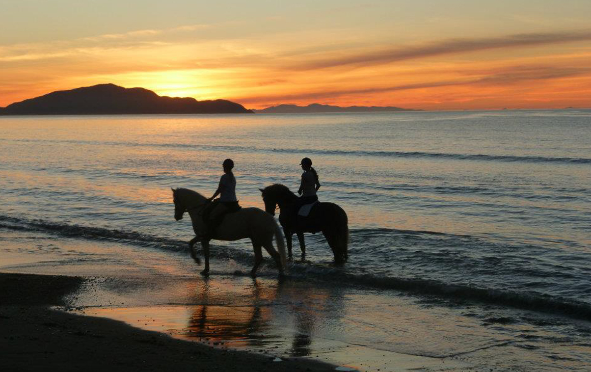 Two people ride horseback along Otaki beach at sunset.