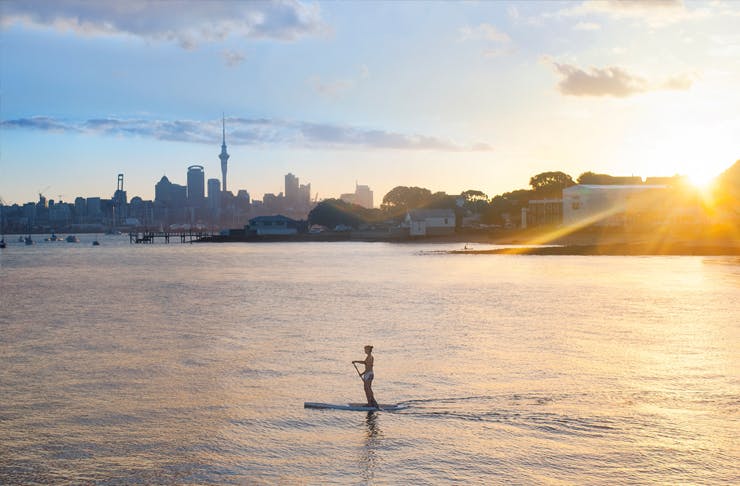 A person paddles across Auckland's Waitemata Harbour on a paddle board at sunrise with the city in the distance.