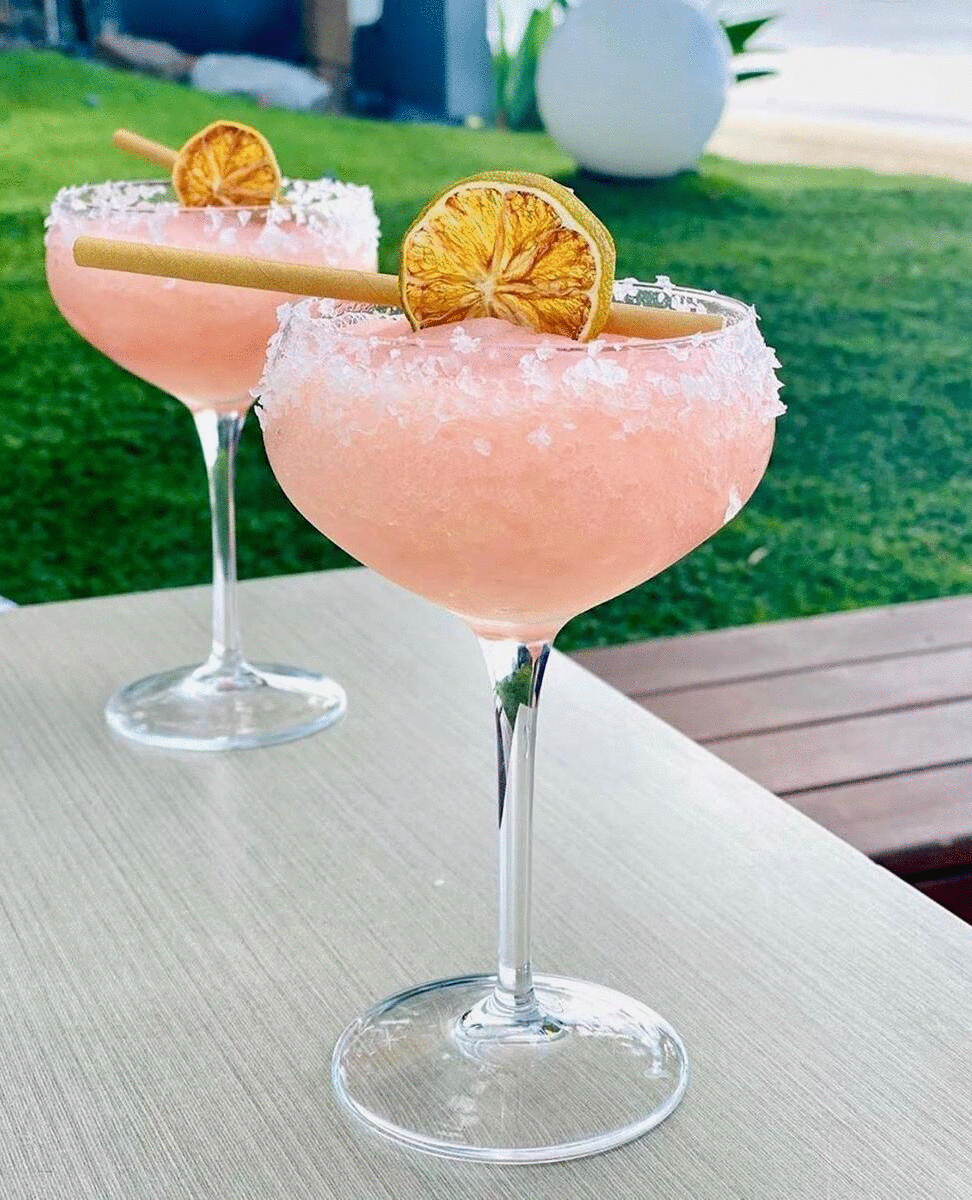 Two pink cocktails sit on a table with the ocean in the background. 