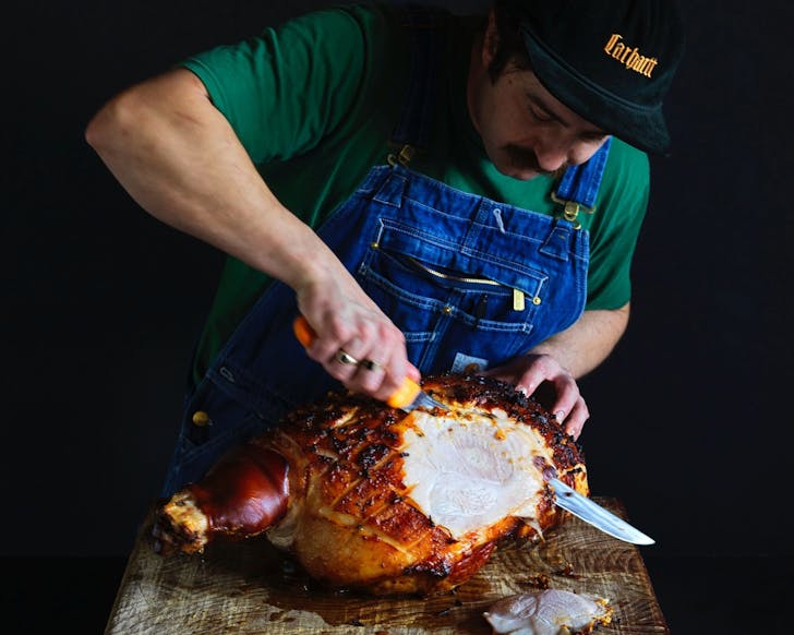A mid-close up picture of a butcher learning over to thinly slice a large piece of meat.