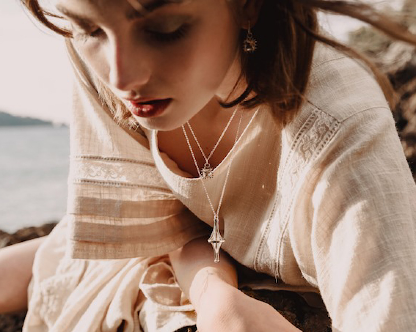 A person wears a linen off-white kaftan and leans over to look into a rock pool. Two Wicca-inspired, crystal necklaces - one small square and one larger diamond - hang around her neck, and there’s a gem dangling from her ear. 