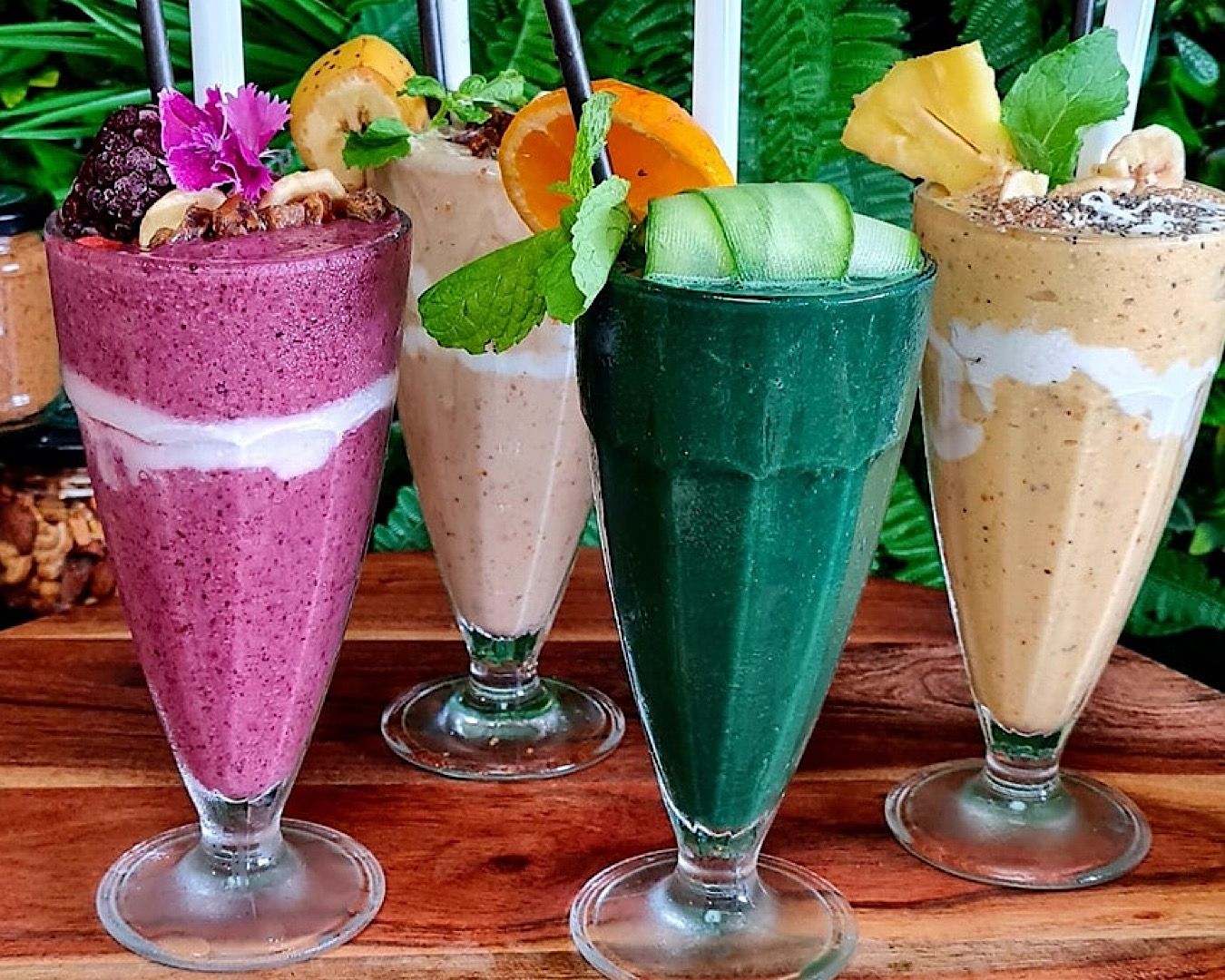 Four vibrant smoothies in old school milkshake glasses: a purple one with gorgeous berry grit, topped with frozen berries and a purple flower, a green one topped with rolled cucumber and a sprig of mint, and two beige-coloured ones topped with fruit and seeds. 