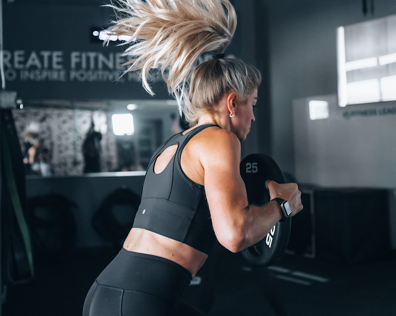 A muscular woman is mid-squat holding a weight plate, her ponytail is flying above her head from momentum. 