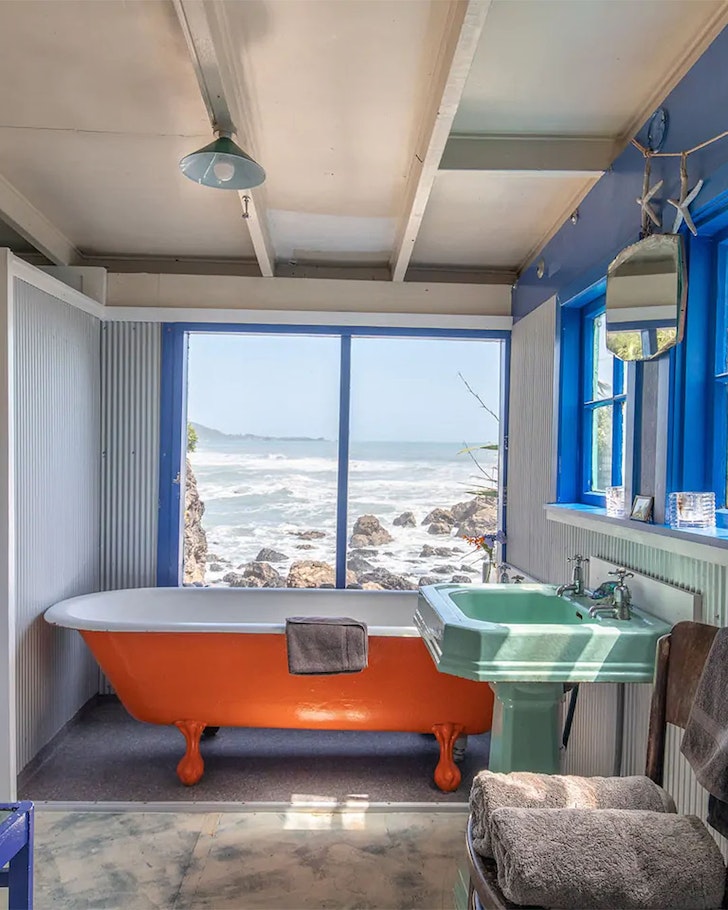 A bathtub sits in the corner of a bach and, outside the window, the ocean laps.