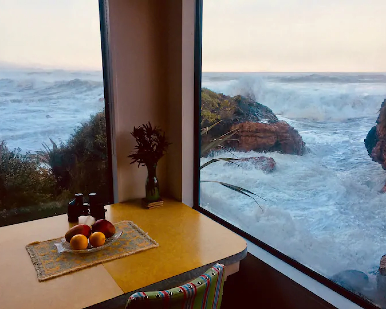 A dining table sits in the corner of a bach and, outside the window, the ocean laps.