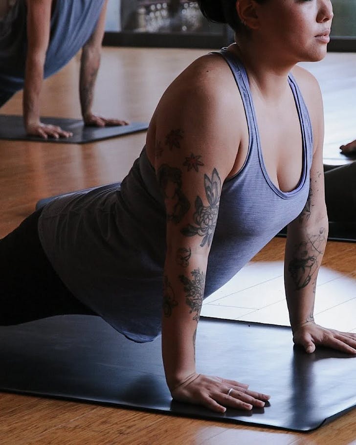 A woman with tattooed arms wearing a blue singlet holds an upward dog pose on a yoga mat.