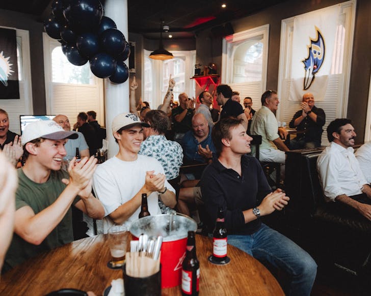 A wide shot of many people sitting at pub tables while smiling and cheering as they look at a screen that is beyond and behind the camera.