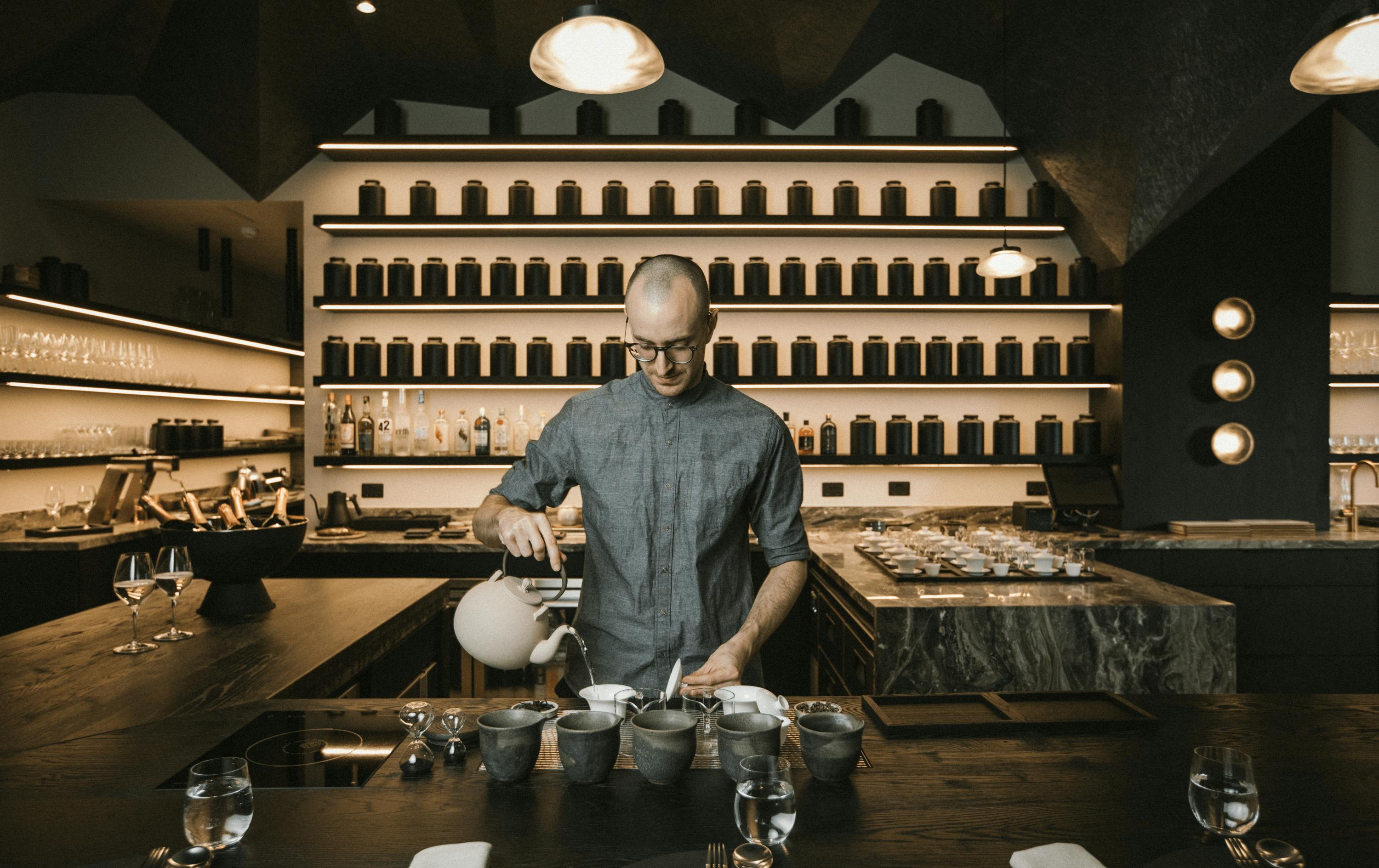 A man standing pouring tea out of a kettle into a cup in a grey shirt.
