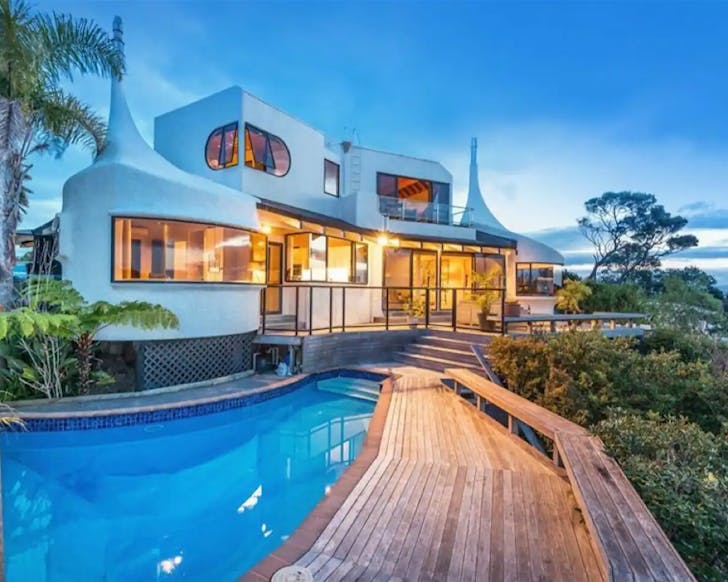â¨â¨A white house with a unique curved design and a curved swimming pool and wooden deck.
