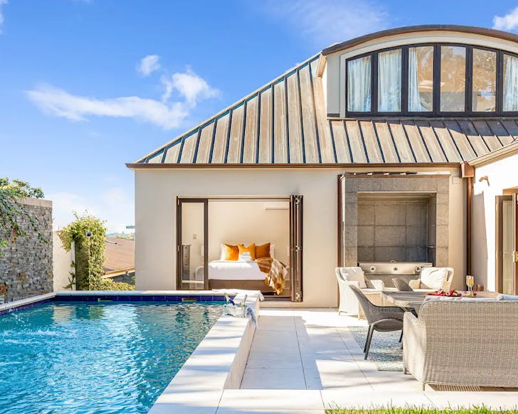 A modern home with a metal roof, curved dormer window and open doors leading to a bedroom and in the foreground a modern pool with a water feature.