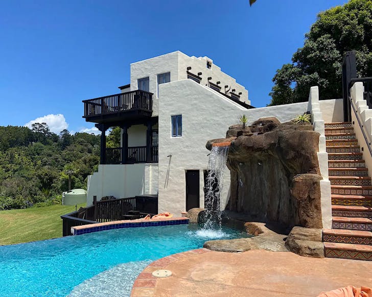 A white Santa Fe-style house with a tiered design, two balconies, and a rock waterfall feature above a curved pool.