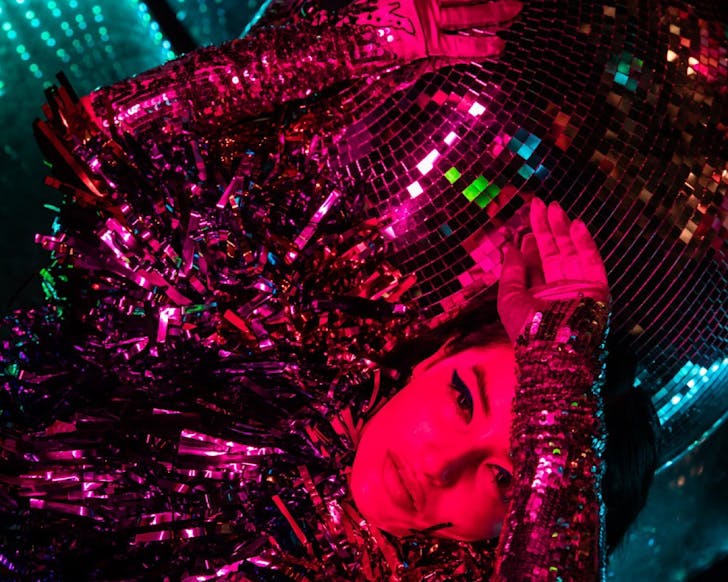 A person with a disco ball. The person is wearing a sequined outfit and has their hand on their head. The disco ball is reflecting light onto the person's face.