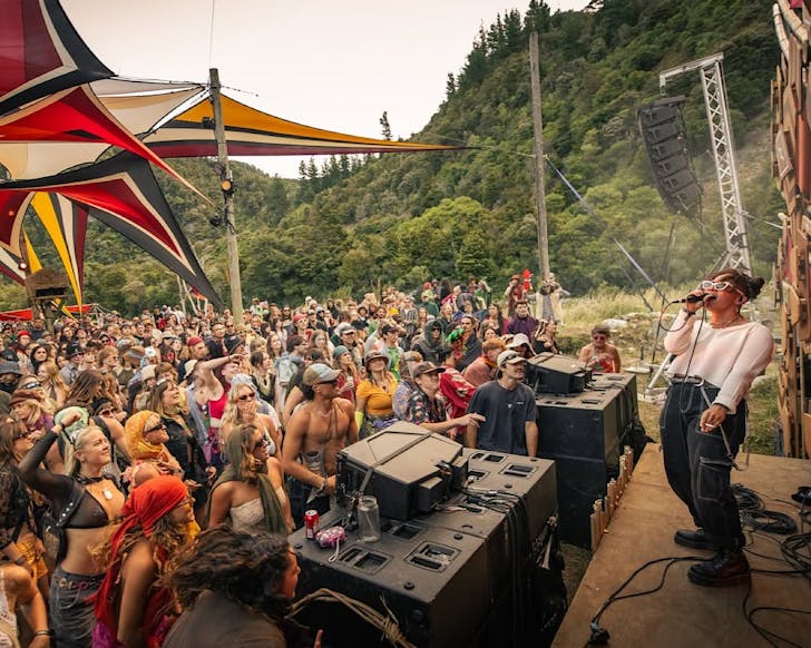 Twisted Frequency festival-goers, under colourful outdoor shade installations, enjoy a woman onstage with a microphone with a backdrop of lush green hills.