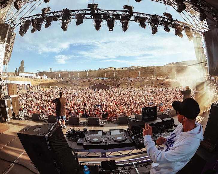 A DJ and two other artists perform in front of a large crowd in the sun at Rolling Meadows.