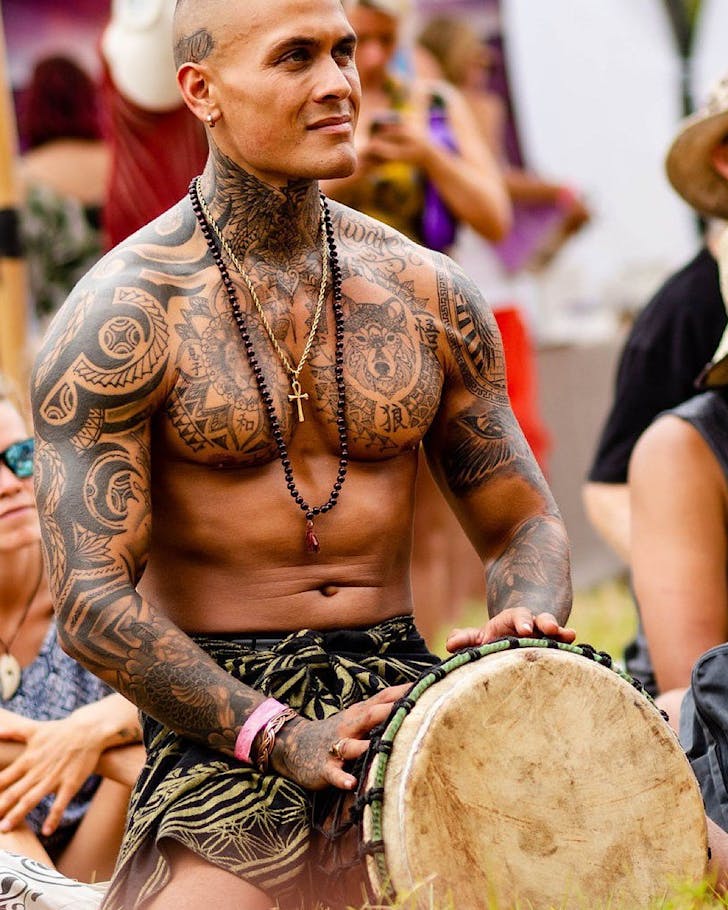 A topless man with traditional tattoos plays a large traditional drum.