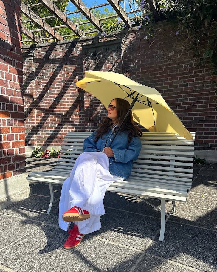 A woman sits on a bench in the sun with a pale yellow BLUNT umbrella to shade her.
