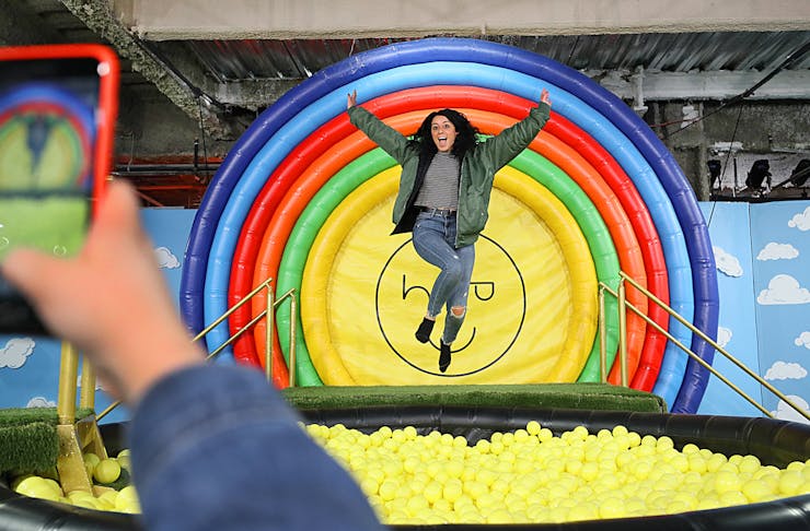 A woman mid-air jumping into a ball pit filled with yellow balls backed by a circular rainbow backdrop.