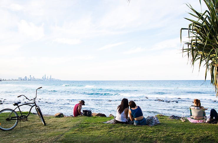 Two young women sit on a picnic rug with their backs to us and are leaning in looking at something on their phones. They overlook the beach at Burleigh Heads, with a Gold Coast cityscape in the distance. 