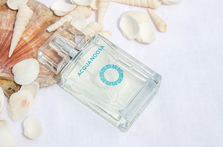 A close-up of a bottle of perfume that says 'Acqua Noosa' among scattered sea shells.