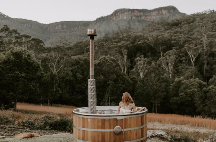 A person in an outdoor hot tub with bushland and mountains in the background. 