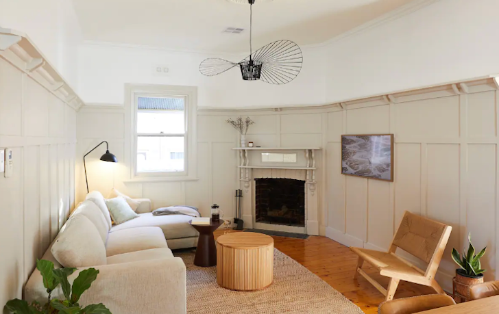 A fireplace sits in the corner of this Airbnb Melbourne living room, opposite a cream-white coloured three-person sofa and a small black lamp handing above.